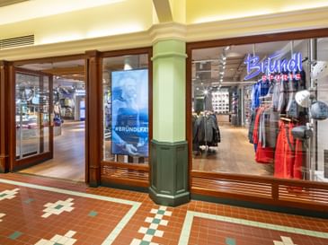 The image shows the glass storefront of the Bründl Sports McArthurGlen Designer Outlet Salzburg shop with a visible “Bründl Sports” logo in the window. Sports clothing, skis, helmets, and accessories are visible inside; a screen shows the text “#BRÜNDLFAN,” and “kassa” can also be seen in the background.