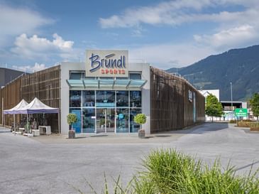 Außenansicht der Bründl Sports Filiale in Saalfelden. Vor dem modernen Geschäftsgebäude mit großem Bründl Sports Logo stehen mehrere Fahrräder unter zwei weißen Zelten. Im Hintergrund sind Berge, Bäume und ein teilweise bewölkter Himmel zu sehen. Sichtbarer Text: „Bründl Sports“ und „Einfahrt“.
