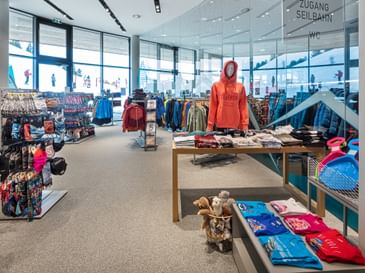 Das Bild zeigt eine Verkaufsfläche eines Bründl Sports Shops mit einer Auswahl an Wintersportbekleidung, Accessoires und Souvenirs. Im Vordergrund steht eine Puppe mit einem Hoodie, daneben sind T-Shirts und weitere Kleidungsstücke ausgestellt. Große Fenster bieten einen Blick nach draußen auf die winterliche Landschaft.