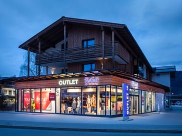 Das Bild zeigt das Bründl Sports Outlet bei Dämmerung. Das Gebäude ist beleuchtet, was die "Outlet" und "Bründl Sports" Schilder gut sichtbar macht. Das Schaufenster zeigt sportliche Winterbekleidung.