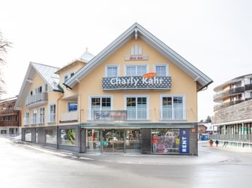 Das Bild zeigt ein Gebäude mit dem "Bründl Sports" Shop im Erdgeschoss und einem Schild für "Charly Kahr" im oberen Bereich, umgeben von anderen Gebäuden in einer winterlichen Umgebung.