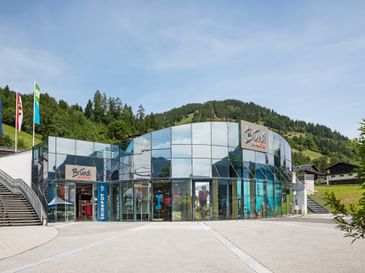 Zu sehen ist der Bründl Sports AreitXpress Talstation Shop mit großer Glasfassade, Shop-Eingang und sichtbarem „Bründl Sports“ Schriftzug an der Fassade. Links befinden sich Fahnen und eine Treppe, im Hintergrund sind Wald, Wiesen und ein Berg zu sehen; am Gebäude steht außerdem der Text „SKIDEPOT“.