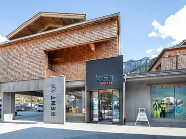 Das Bild zeigt den Eingangsbereich eines Bründl Sports Shops in einem modernen Gebäude mit Holzverkleidung und Glasfronten, umgeben von einer alpinen Landschaft.