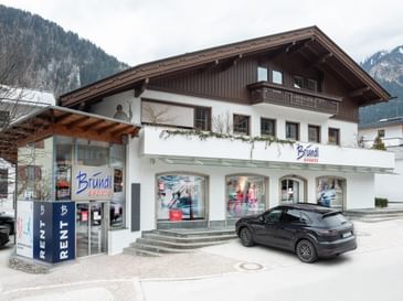 Das Bild zeigt einen Bründl Sports Shop in einem alpenländischen Gebäude mit Schaufenstern, einem Auto davor und Bergen im Hintergrund.