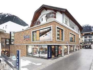 Bründl Sports Ischgl Zentrum in daylight, featuring a stone façade, large display windows and Bründl Sports logos. A “RENT” sign stands outside the store, with patches of snow along the street and snow-covered mountains in the background.