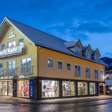 Das Bild zeigt ein Gebäude mit dem "Bründl Sports" Shop im Erdgeschoss und einem Schild für "Charly Kahr" im oberen Bereich, umgeben von anderen Gebäuden in einer winterlichen Umgebung.