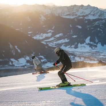 Two skiers descend a snowy ski slope in the low evening sun. A snow-covered mountain panorama stretches across the background.