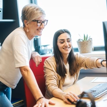 One woman is sitting at a computer pointing at the screen, while another woman stands beside her smiling. On the desk are a monitor, keyboard, and mouse, and in the background there are folders and a plant.