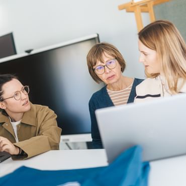 Three women are standing and sitting at a table with open laptops, talking to each other. One woman looks at the others while an older woman in the middle speaks and the third listens attentively.