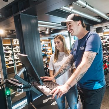 A Bründl Sports employee is carrying out a bike fitting using a digital station while a customer stands beside him and smiles. Bicycles, e-bikes and sports accessories are visible in the shop background; the Bründl Sports logo can be seen on the shirt and a bag.