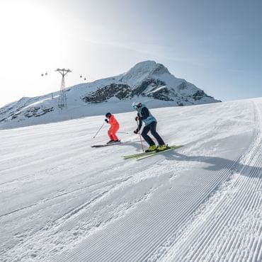 Das Bild zeigt zwei Skifahrer, die nebeneinander eine frisch präparierte Piste hinunterfahren. Einer trägt eine orangefarbene Skiausrüstung, der andere eine blaue. Im Hintergrund sind schneebedeckte Berge und ein Mast einer Seilbahn zu sehen, die die alpine Landschaft abrunden.