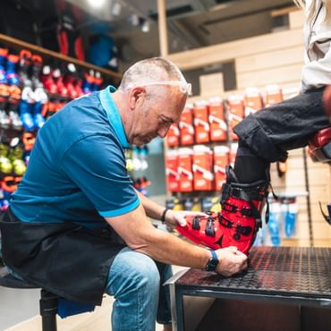 Ein Bründl Sports Mitarbeiter passt einer Kundin im Shop einen roten Skischuh an. Er sitzt vor einer Wand mit Skischuhen und sorgt für die perfekte Passform und optimalen Komfort.