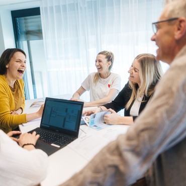 Five people are sitting around a table in a bright room, laughing together. A laptop and several documents are on the table, being looked at by the group.