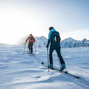 Two people are ascending a snowy mountain landscape in ski touring gear, with the sun shining behind the peaks.