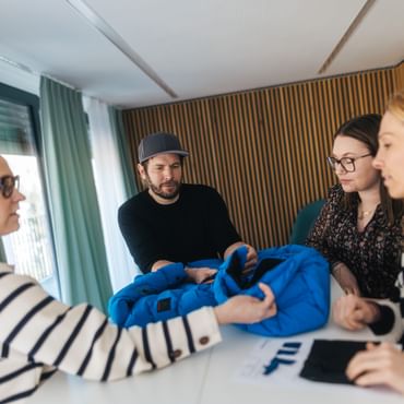Four people stand around a table examining a blue jacket together. They hold the garment and look at it while discussing it.