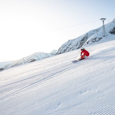 Das Bild zeigt eine Skifahrerin in roter Kleidung, die mit hoher Geschwindigkeit eine frisch präparierte Piste hinunterfährt. Der Schnee wirbelt hinter ihr auf, und im Hintergrund sind hohe Berge zu sehen.