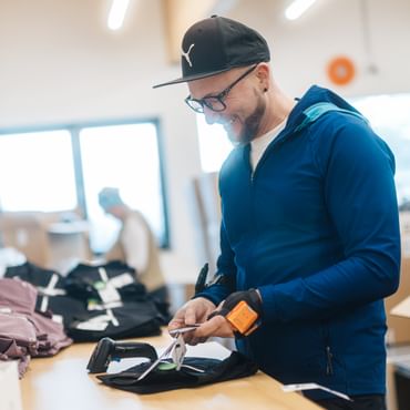 A man wearing a cap and glasses stands at a table attaching a label to a piece of clothing. Other clothing items are beside him, and boxes and another person working can be seen in the background.