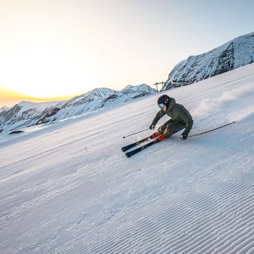Das Bild zeigt einen Skifahrer, der bei Sonnenaufgang eine schneebedeckte Piste hinunterfährt, umgeben von Berglandschaft.