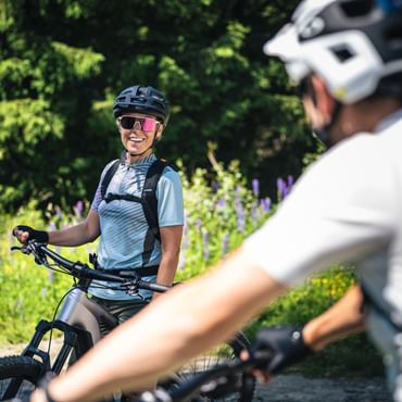 A person wearing a helmet, sunglasses, and a backpack sits on a mountain bike and smiles toward another person who is blurred in the foreground. In the background, dense green trees and blooming plants line a forest trail.