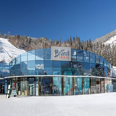 Zu sehen ist der Bründl Sports AreitXpress Talstation Shop mit großer blauer Glasfassade direkt an einer verschneiten Skipiste. Auf dem Gebäude sind der „Bründl Sports“ Schriftzug sowie die Texte „SKIDEPOT“, „SHOP“, „RENT“, „DEPOT“ und „SERVICE“ sichtbar; im Hintergrund sind Skilift, Wald und verschneite Berge zu sehen.