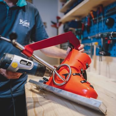 A person is heating a bright orange ski boot shell in a workshop using a heat gun directed at the outer side of the boot. The ski boot is fixed on a metal device, and tools are visible on a blue wall in the background.