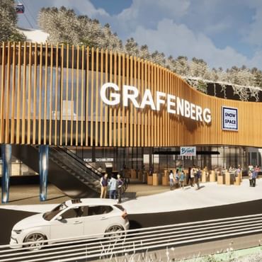 Ein modernes Gebäude mit geschwungener Holzfassade und dem Schriftzug „GRAFENBERG“ steht vor verschneiten Bäumen. Vor dem Eingang stehen mehrere Menschen, ein Auto fährt vorbei, und Schilder mit „Flachau Snow Space Salzburg“, „Bründl Sports“ sowie „Ticketschalter“ sind sichtbar, während oben eine Gondelbahn verläuft.
