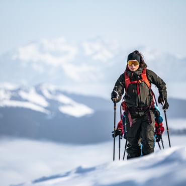 Eine Person in Skitouren-Ausrüstung steigt einen schneebedeckten Hang hinauf, während im Hintergrund eine beeindruckende Berglandschaft mit wolkenverhangenen Tälern zu sehen ist.