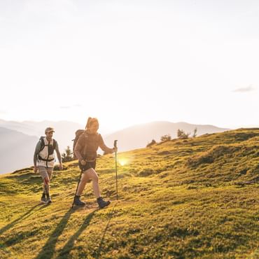 Zwei Personen mit Rucksäcken wandern über einen sanft ansteigenden, grasbewachsenen Hügel. Die tief stehende Sonne wirft lange Schatten auf die Wiese, im Hintergrund sind Berge im warmen Abendlicht zu sehen.