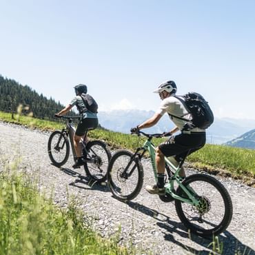 Two people wearing helmets and backpacks ride mountain bikes one behind the other along an uphill gravel trail. A fence runs alongside the path, and green hills and mountains can be seen in the background under a clear blue sky.