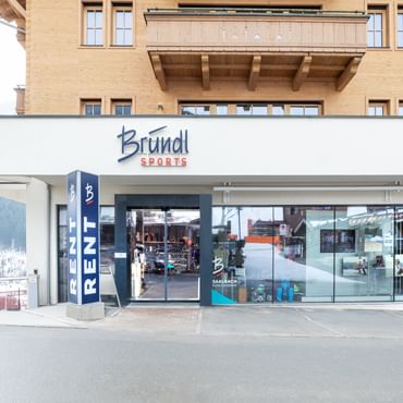 Das Bild zeigt den Eingang eines Bründl Sports Shops in einem alpinen Gebäude. Über dem Eingang befindet sich das Logo, und der Schriftzug "RENT" weist auf die Verleihmöglichkeit hin.