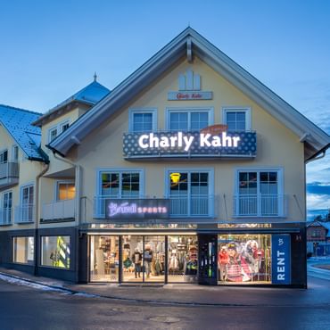 Das Bild zeigt ein Gebäude mit dem "Bründl Sports" Shop im Erdgeschoss und einem Schild für "Charly Kahr" im oberen Bereich, umgeben von anderen Gebäuden in einer winterlichen Umgebung.