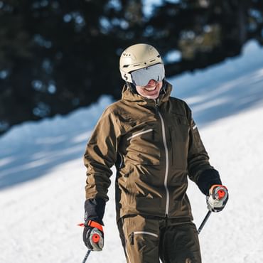 Ein lächelnder Skifahrer in brauner Skibekleidung steht mit Helm, Skibrille und Skistöcken auf einer verschneiten Skipiste. Im Hintergrund ist ein dunkler Wald zu sehen.