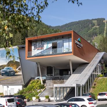 Das Bild zeigt ein Gebäude mit großer Glasfassade und markantem, schrägem Dach. Vor dem Eingang sind Autos zu sehen. Auf der Seite befinden sich das Bründl Sports-Logo und das Schmitten-Logo auf der braunen Fassade. Die Umgebung ist von dichten Wäldern umgeben, was eine idyllische Kulisse bildet.
