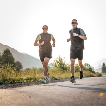 Zwei Läufer joggen nebeneinander auf einem asphaltierten Weg durch eine alpine Landschaft bei tief stehender Sonne.
