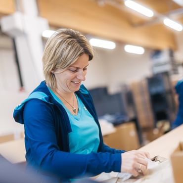 A woman stands at a worktable placing an item into a bag or packaging. In the background, there are boxes and another person working.