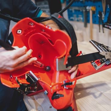 A person is working on a bright red ski boot shell fixed on a workbench in a workshop. Various tools and a blue tool wall are visible in the background.