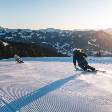 Zwei Skifahrer fahren mit sportlichen Skischwüngen über eine präparierte Skipiste. Die tief stehende Sonne beleuchtet die schneebedeckte Berglandschaft im Morgenlicht.