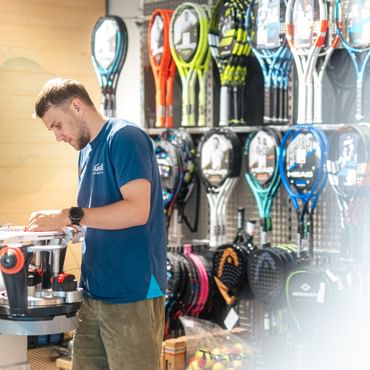 An employee stands at a stringing machine, working intently on a tennis racket. In the background, numerous rackets are displayed on a wall, sorted by brand and model.<br/>