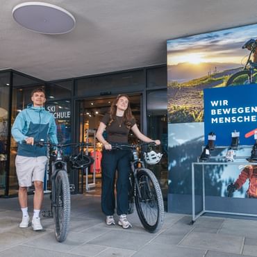 Ein junger Mann und eine junge Frau stehen mit Mountainbikes vor einem Sportgeschäft. Im Hintergrund sind Kleidung, Fahrradhelme und ein großes Werbeplakat mit einem Fahrradfahrer zu sehen. Auf dem blauen Schild steht der Text „WIR BEWEGEN MENSCHEN“.