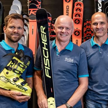 Das Bild zeigt Bründl Sports Mitarbeiter, die vor einer Wand mit verschiedenen Skiern stehen. Sie tragen alle blaue Polohemden mit dem Bründl Sports-Logo. Der Mann links hält ein Paar gelbe Fischer-Skischuhe, der Mann in der Mitte hält einen Fischer-Ski, und der Mann rechts hält einen Atomic-Ski. Alle drei lächeln in die Kamera. Im Hintergrund sind weitere Skier verschiedener Marken wie Fischer, Rossignol und Atomic zu sehen.