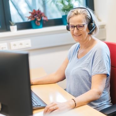A woman wearing glasses and a headset sits at a desk looking at a computer screen. She holds a mouse in her hand, with a keyboard on the desk and plants visible by the window in the background.