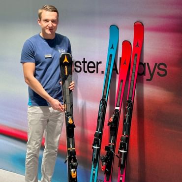 A Bründl Sports employee stands in a modern showroom in front of a wall with the Atomic logo, holding a black ski with orange details. Next to him are three more Atomic skis in blue, pink, and black.