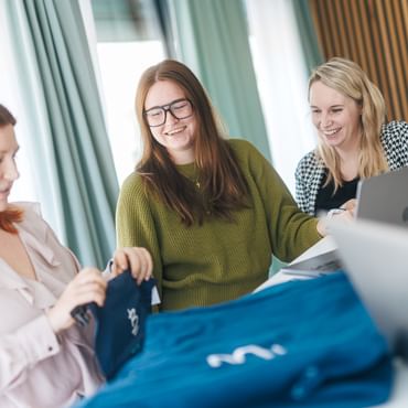 Drei Frauen stehen an einem Tisch, auf dem blaue Kleidungsstücke liegen, und betrachten diese gemeinsam. Eine Frau hält ein Kleidungsstück in der Hand, während die anderen lächeln und darauf schauen; im Hintergrund sind Laptops zu sehen.