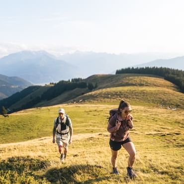 A woman wearing a backpack hikes up a grassy hill, while a man with a cap and backpack follows a few steps behind. In the background, rolling hills, scattered trees, and distant mountains can be seen under a clear sky.