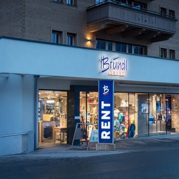 Das Bild zeigt den Eingang eines Bründl Sports Shops in einem alpinen Gebäude. Über dem Eingang befindet sich das Logo, und der Schriftzug "RENT" weist auf die Verleihmöglichkeit hin.