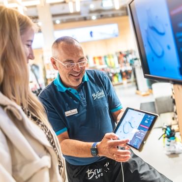 A Bründl Sports employee advises a customer in the shop, showing a digital ski boot analysis on a tablet. Sports equipment and ski gear are visible in the background.