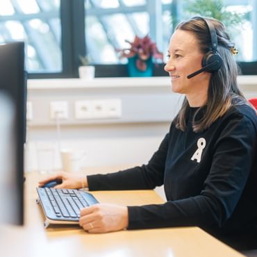 A woman sits on a red office chair at a desk wearing a headset while using a keyboard and mouse. She looks at a monitor, with windows and plants visible in the background.