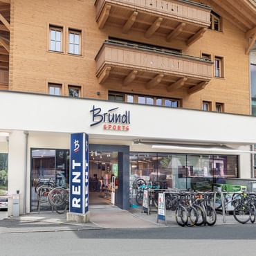 Das Bild zeigt die Außenansicht eines Bründl Sports Shops in einem alpinen Gebiet. Vor dem Geschäft stehen mehrere Fahrräder und E-Bikes zur Miete, gekennzeichnet durch ein großes "RENT"-Schild. Das Gebäude hat eine Holzverkleidung und Balkone, typisch für die Region. Durch die Schaufenster sind weitere Fahrräder und Sportausrüstung im Inneren sichtbar. Im Hintergrund erstrecken sich grüne Berge.