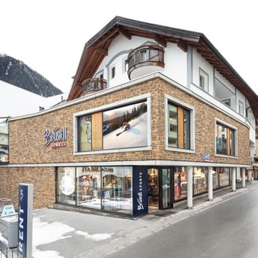 Bründl Sports Ischgl Zentrum mit Steinfassade, großen Schaufenstern und Bründl Sports Logos bei Tageslicht. Vor dem Shop steht ein „RENT“-Schild; Schneereste säumen die Straße und schneebedeckte Berge liegen im Hintergrund.