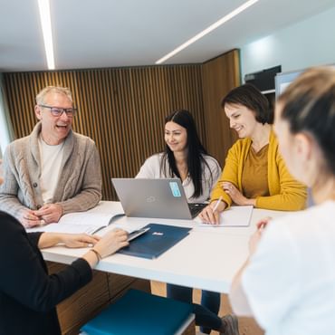 Fünf Menschen vom Personalteam stehen an einem Tisch in einem modernen Raum und unterhalten sich. In der Mitte steht ein Laptop, daneben liegen Dokumente und ein Notizbuch, während einige der Personen lächeln und schreiben.